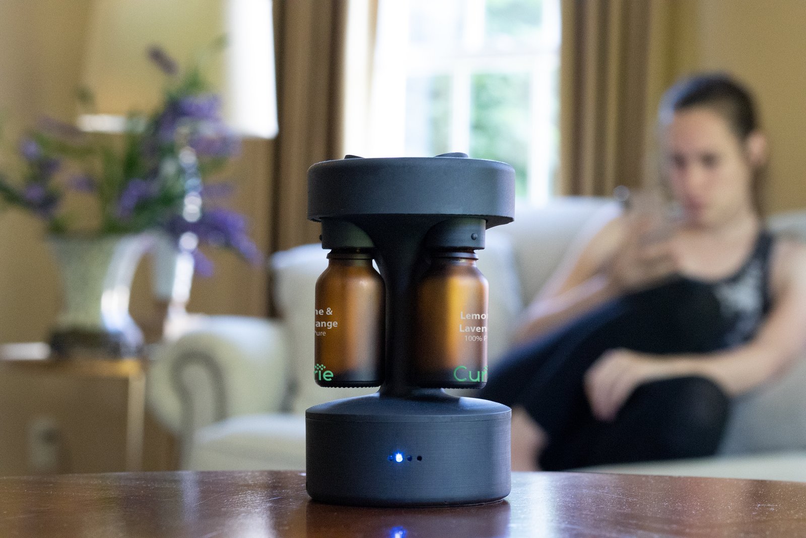 Curie diffuser on a living-room table, amber bottles loaded, blue LED lit, a person seated in the background.
